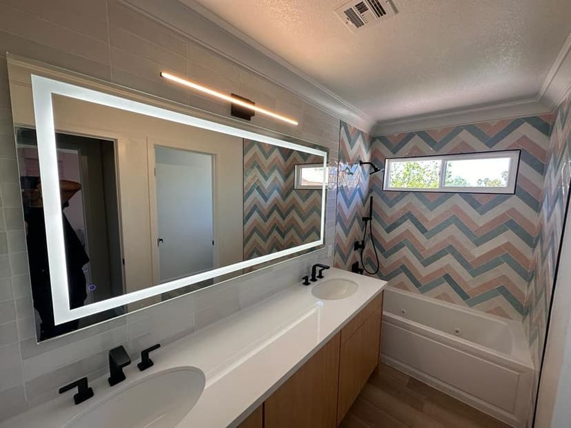 Modern bathroom with a large illuminated mirror, double sinks, and chevron-patterned wall.